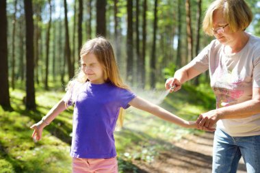 Middle age woman applying insect repellent to her granddaughter before forest hike beautiful summer day. Protecting children from biting insects at summer. Active leisure with kids. clipart