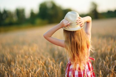 Sevimli kız mutlu bir şekilde yürürken buğday alanında sıcak ve güneşli yaz akşamı hasır şapka giyiyor. Sevimli küçük çocuk çavdar alanında günbatımı.