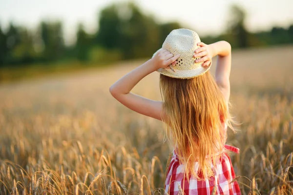 Sevimli kız mutlu bir şekilde yürürken buğday alanında sıcak ve güneşli yaz akşamı hasır şapka giyiyor. Sevimli küçük çocuk çavdar alanında günbatımı.