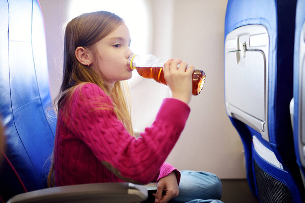 Adorable little girl traveling by an airplane. Child sitting by aircraft window and drinking a beverage. Traveling with kids abroad.