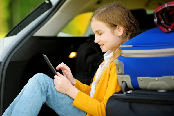 愛らしい小さな女の子は彼女の両親と一緒に旅行に行く準備ができて 子供を車のトランクに座っていると彼女の Ebook を読んでします 子供たちと車で旅行 — ストック写真