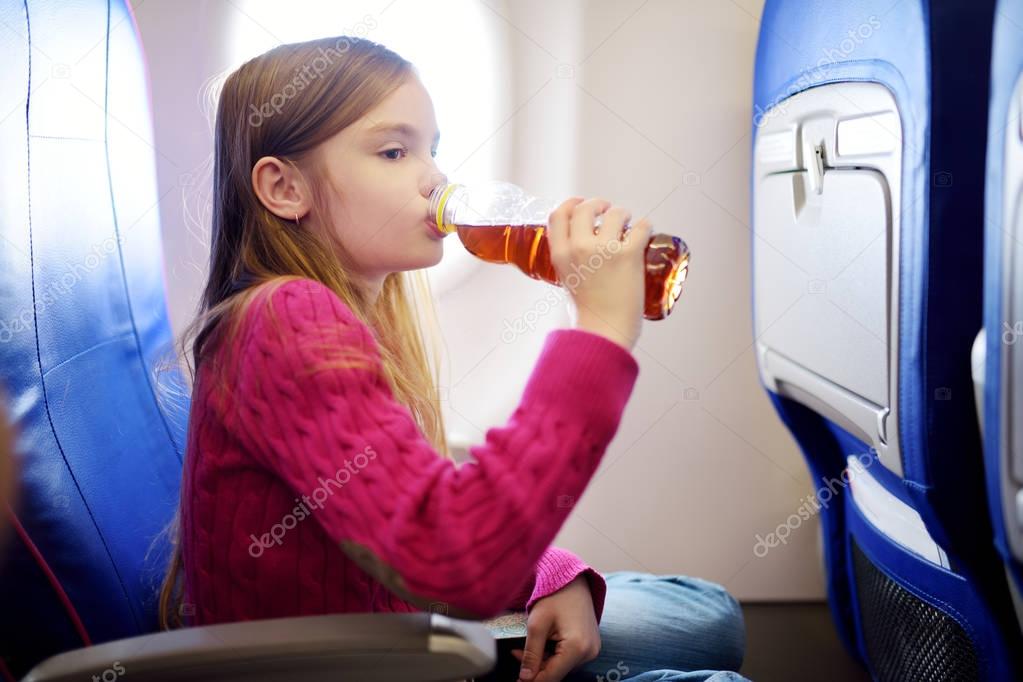 Adorable little girl traveling by an airplane. Child sitting by aircraft window and drinking a beverage. Traveling with kids abroad.