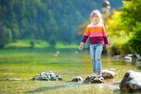 Adorable girl playing by Konigssee lake in Germany on warm summer day. Cute child having fun feeding ducks and throwing stones into the lake. Summer activities for kids.