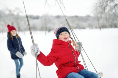 İki sevimli genç kız güzel kış parkında salıncakta birlikte eğleniyorlar. Karda oynayan sevimli kız kardeşler.