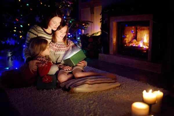Felice giovane madre e le sue due piccole figlie aprendo un magico regalo di Natale da un camino in un accogliente salotto buio la vigilia di Natale . — Foto Stock