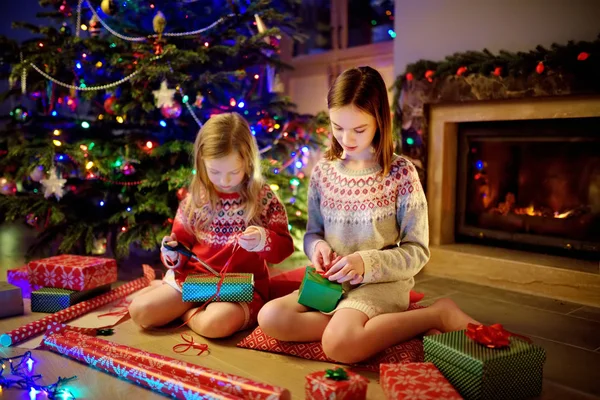 Jolies jeunes sœurs enveloppant des cadeaux ensemble par une cheminée dans un salon sombre confortable la veille de Noël. Célébrer Noël à la maison . — Photo