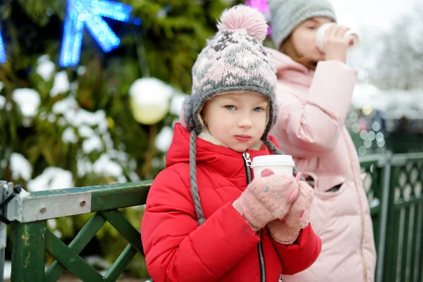 Две очаровательные сестры пьют горячий шоколад на традиционной рождественской ярмарке в Риге, Латвия. Дети наслаждаются сладостями, сладостями и пряниками на рождественском рынке . — стоковое фото