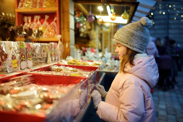 ラトビアのリガにある伝統的なクリスマスマーケットでお菓子を選ぶかわいい女の子。Xmasでキャンディとクッキーを買う子供. — ストック写真