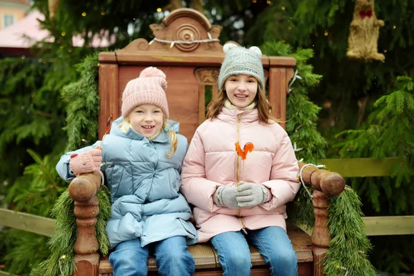 Dwie urocze siostry posiadające lizaki w kształcie koguta na tradycyjnym jarmarku bożonarodzeniowym w Rydze na Łotwie. Dzieci delektujące się słodyczami, cukierkami i piernikiem na rynku świątecznym. — Zdjęcie stockowe