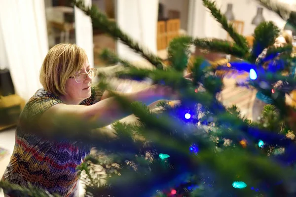 Starší žena zdobí vánoční stromek zlatým věncem. Ořezávání vánočního stromečku. — Stock fotografie