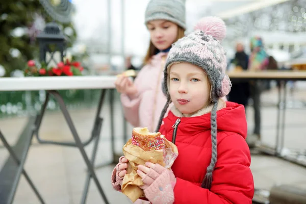 Deux jolies jeunes sœurs mangeant du trdelnik tchèque lors de la traditionnelle foire de Noël à Vilnius, en Lituanie. Enfants dégustant des bonbons, des bonbons et du pain d'épice sur le marché de Noël . — Photo