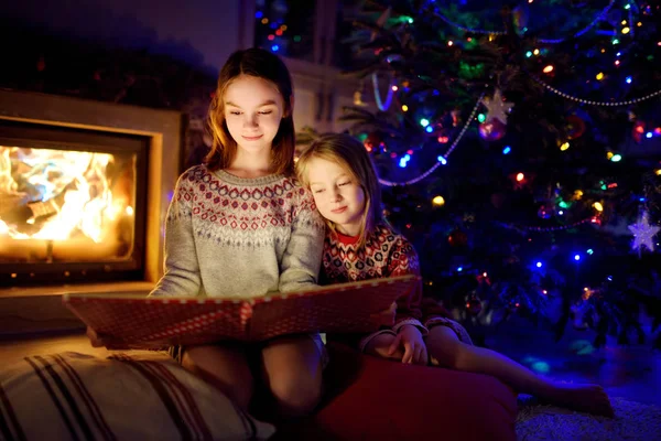 Happy young sisters reading a story book together by a fireplace in a cozy dark living room on Christmas eve. Celebrating Xmas at home. — Stock Photo, Image