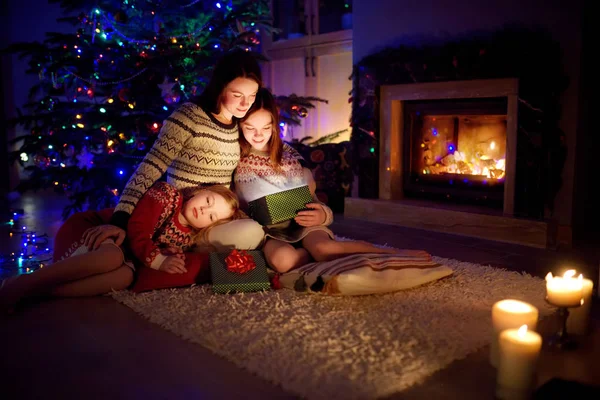 Happy young mother and her two small daughters opening a magical Christmas gift by a fireplace in a cozy dark living room on Christmas eve. — Stock Photo, Image