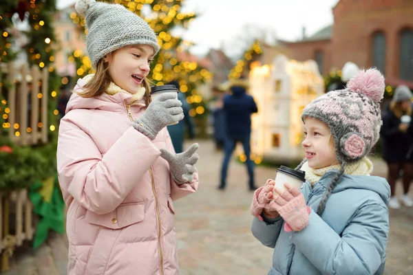 ラトビアのリガで伝統的なクリスマスフェアでホットチョコレートを飲む2人の愛らしい姉妹。クリスマスマーケットでお菓子やキャンディー、ジンジャーブレッドを楽しむ子供たち. — ストック写真