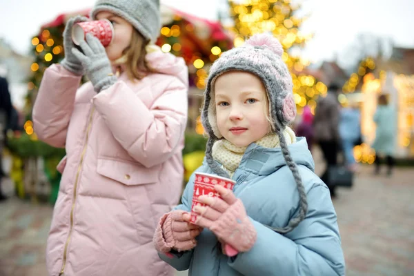 ラトビアのリガで伝統的なクリスマスフェアでホットチョコレートを飲む2人の愛らしい姉妹。クリスマスマーケットでお菓子やキャンディー、ジンジャーブレッドを楽しむ子供たち. — ストック写真