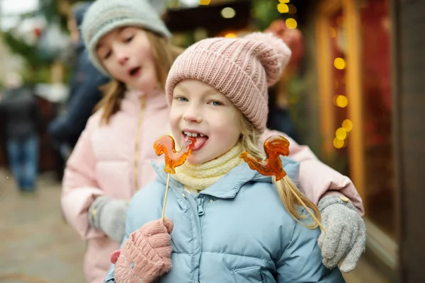 Två bedårande systrar som har tuppformade klubbor på traditionell julmässa i Riga, Lettland. Barn som njuter av sötsaker, godis och pepparkakor på julmarknaden. — Stockfoto