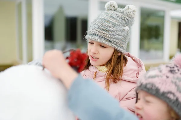 Adorable young girls building a snowman in the backyard. Cute children playing in a snow. Winter activities for kids. — Stock Photo, Image