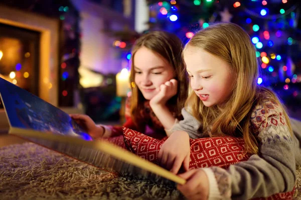 Happy young sisters reading a story book together by a fireplace in a cozy dark living room on Christmas eve. Celebrating Xmas at home. — Stock Photo, Image