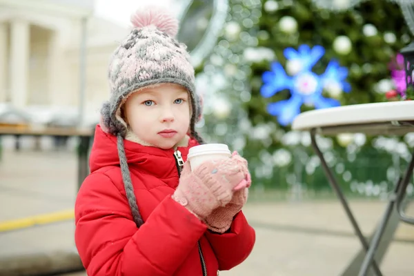 Jolie jeune fille buvant du chocolat chaud sur la foire de Noël traditionnelle à Riga, Lettonie. Enfant dégustant des bonbons, des bonbons et du pain d'épice sur le marché de Noël . — Photo