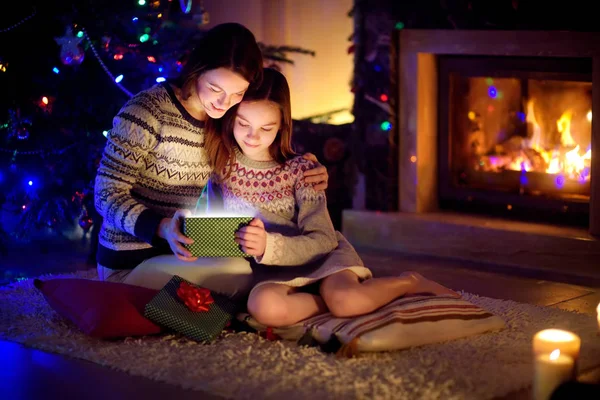 Happy young mother and her young daughter opening a magical Christmas gift by a fireplace in a cozy dark living room on Christmas eve.