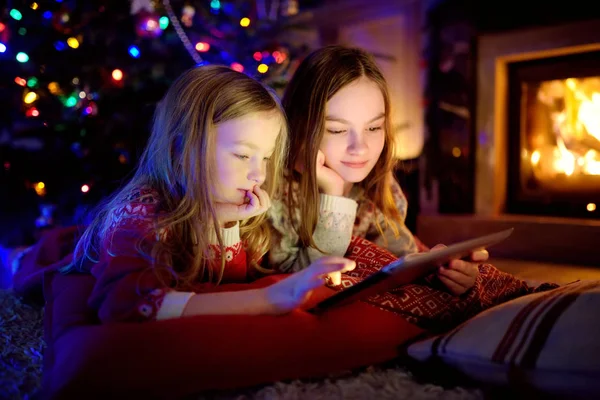 Två söta unga systrar använder en surfplatta hemma vid en öppen spis i varmt och mysigt vardagsrum på julafton. — Stockfoto