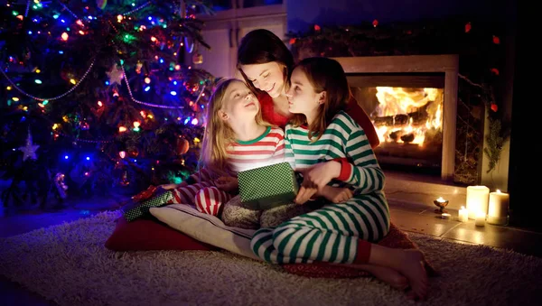 Feliz madre joven y sus dos hijas pequeñas abriendo un regalo mágico de Navidad junto a una chimenea en una acogedora sala de estar oscura en la víspera de Navidad . — Foto de Stock