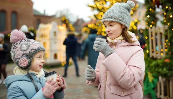 Duas irmãs adoráveis bebendo chocolate quente na tradicional feira de Natal em Riga, Letônia. Crianças desfrutando de doces, doces e pão de gengibre no mercado de Natal . — Fotografia de Stock