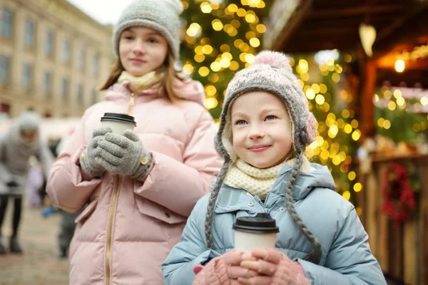Riga, Letonya 'da geleneksel Noel fuarında sıcak çikolata içen iki sevimli kız kardeş. Çocuklar Xmas pazarında tatlı, şeker ve zencefilli ekmek yiyor.. — Stok fotoğraf