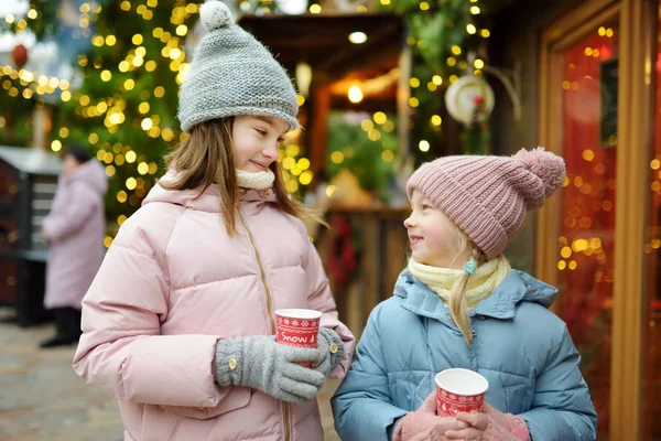 Deux adorables sœurs boivent du chocolat chaud lors de la foire traditionnelle de Noël à Riga, en Lettonie. Enfants dégustant des bonbons, des bonbons et du pain d'épice sur le marché de Noël . — Photo