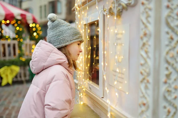 ラトビアのリガにある伝統的なクリスマスマーケットでお菓子を選ぶかわいい女の子。Xmasでキャンディとクッキーを買う子供. — ストック写真