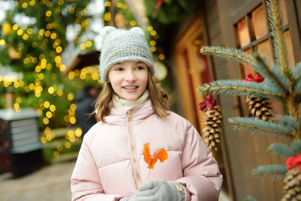 ラトビアのリガで開催される伝統的なクリスマス・フェアで、雄鶏の形をしたロリポップを持つかわいい女の子。クリスマスマーケットでお菓子、キャンディー、ジンジャーブレッドを楽しむ子供. — ストック写真