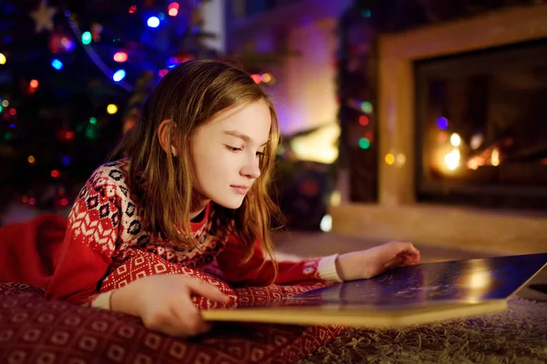 Glückliches junges Mädchen, das an Heiligabend in einem gemütlichen dunklen Wohnzimmer am Kamin ein Märchenbuch liest. Weihnachten zu Hause feiern. — Stockfoto