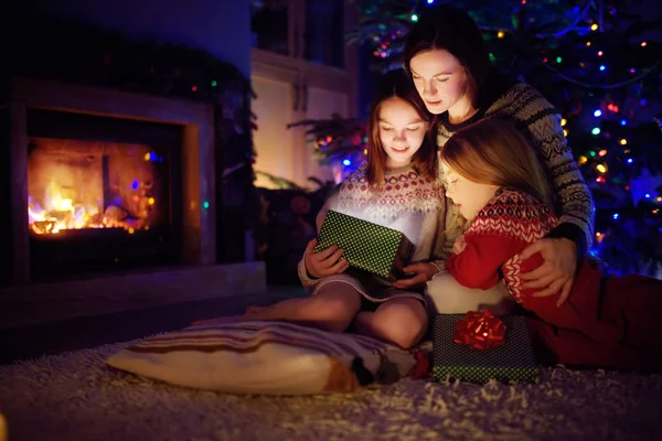 Joyeux jeune mère et ses deux petites filles ouvrant un cadeau de Noël magique près d'une cheminée dans un salon sombre confortable la veille de Noël . — Photo