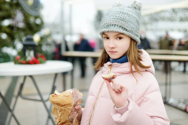 Menina bonito comer trdelnik checo na feira de Natal tradicional em Vilnius, Lituânia. Criança desfrutando de doces, doces e pão de gengibre no mercado de Natal . — Fotografia de Stock