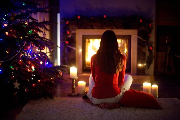 Glückliche junge Frau, die es sich an Heiligabend am Kamin in einem gemütlichen dunklen Wohnzimmer gut gehen lässt. Weihnachten zu Hause feiern. — Stockfoto