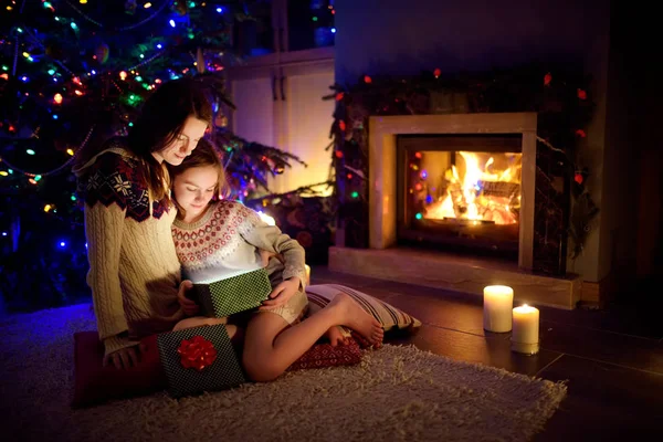 Happy young mother and her young daughter opening a magical Christmas gift by a fireplace in a cozy dark living room on Christmas eve. — Stock Photo, Image