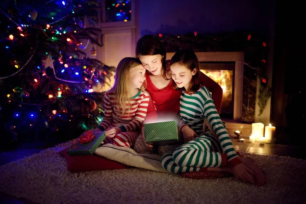 Lycklig ung mor och hennes två små döttrar öppnar en magisk julklapp vid en öppen spis i ett mysigt mörkt vardagsrum på julafton. — Stockfoto