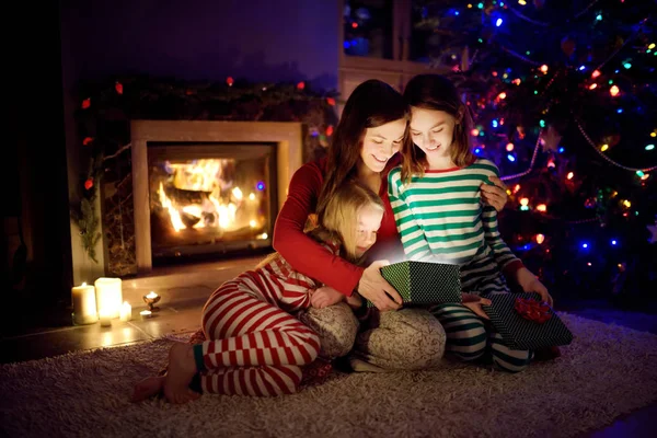 Felice giovane madre e le sue due piccole figlie aprendo un magico regalo di Natale da un camino in un accogliente salotto buio la vigilia di Natale . — Foto Stock