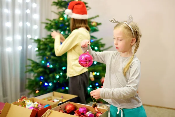 Zwei entzückende Schwestern schmücken zu Hause einen Weihnachtsbaum mit bunten Glaskugeln. Familienfreizeit zur wunderbaren Weihnachtszeit. — Stockfoto