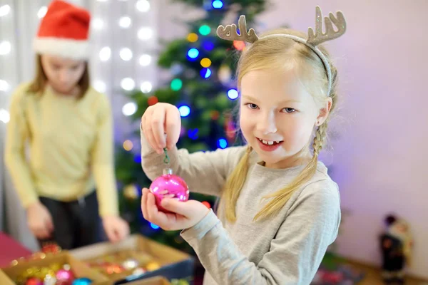 Két imádnivaló nővér, akik egy karácsonyfát díszítenek színes üvegfalatokkal otthon. Családi szabadidő a csodálatos karácsony idején. — Stock Fotó