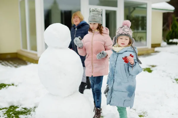 Leuke jonge meisjes en hun oma bouwen een sneeuwpop in de achtertuin. Leuke kinderen spelen in een sneeuw. — Stockfoto