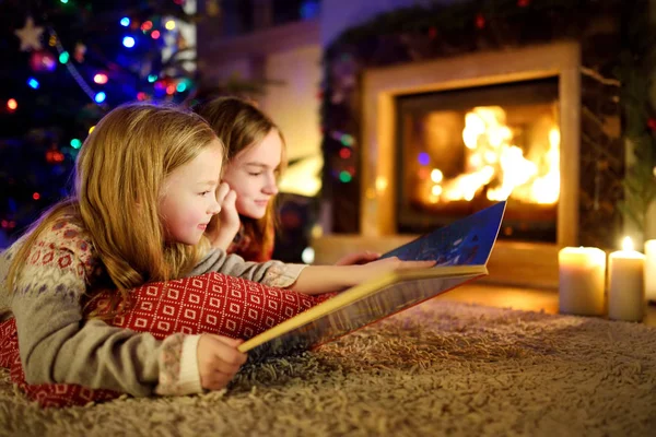 Giovani sorelle felici che leggono insieme un libro di storie accanto a un camino in un accogliente salotto buio la vigilia di Natale. Festeggiare Natale a casa . — Foto Stock