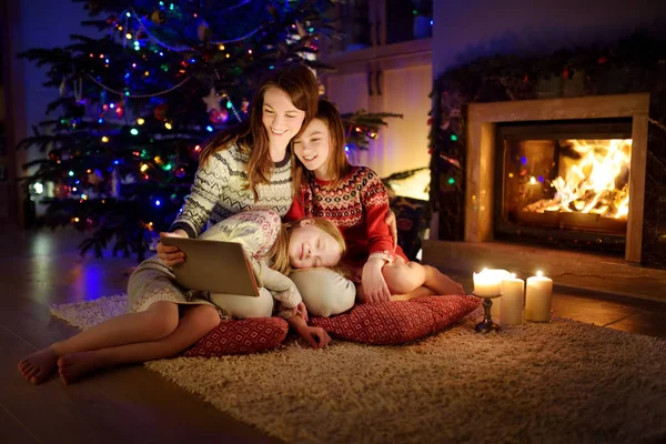 Μητέρα και δύο χαριτωμένες κόρες της χρησιμοποιώντας ένα tablet pc στο σπίτι από ένα τζάκι στο ζεστό και άνετο σαλόνι την παραμονή των Χριστουγέννων. — Φωτογραφία Αρχείου