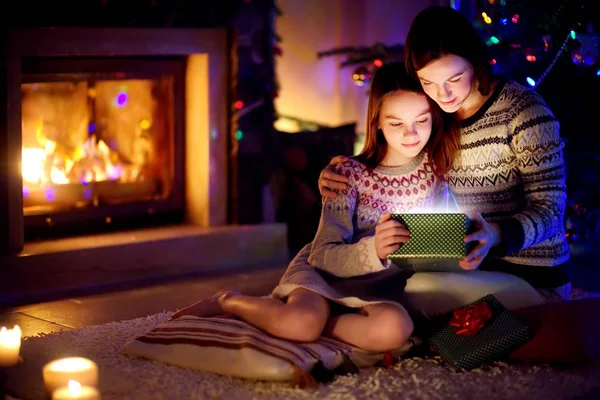 Happy young mother and her young daughter opening a magical Christmas gift by a fireplace in a cozy dark living room on Christmas eve. — Stock Photo, Image