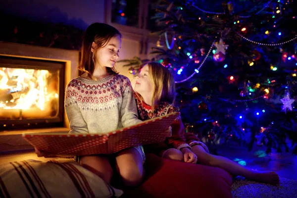 Happy young sisters reading a story book together by a fireplace in a cozy dark living room on Christmas eve. Celebrating Xmas at home. — Stock Photo, Image