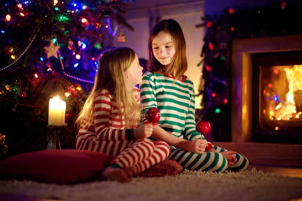 Irmãs felizes comendo maçãs vermelhas cobertas com açúcar gelado junto a uma lareira em uma acolhedora sala de estar escura na véspera de Natal. Celebrando o Natal em casa . — Fotografia de Stock