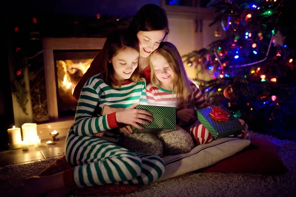 Happy young mother and her two small daughters opening a magical Christmas gift by a fireplace in a cozy dark living room on Christmas eve.