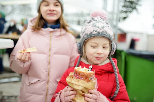 リトアニアのヴィリニュスで伝統的なクリスマスフェアでチェコのトルデリクを食べる2人のかわいい妹。クリスマスマーケットでお菓子やキャンディー、ジンジャーブレッドを楽しむ子供たち. — ストック写真