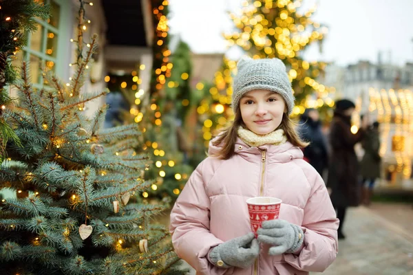 ラトビアのリガで開催される伝統的なクリスマスフェアでホットチョコレートを飲むかわいい女の子。クリスマスマーケットでお菓子、キャンディー、ジンジャーブレッドを楽しむ子供. — ストック写真