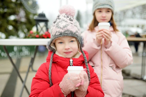 Duas irmãs adoráveis bebendo chocolate quente na tradicional feira de Natal em Riga, Letônia. Crianças desfrutando de doces, doces e pão de gengibre no mercado de Natal . — Fotografia de Stock